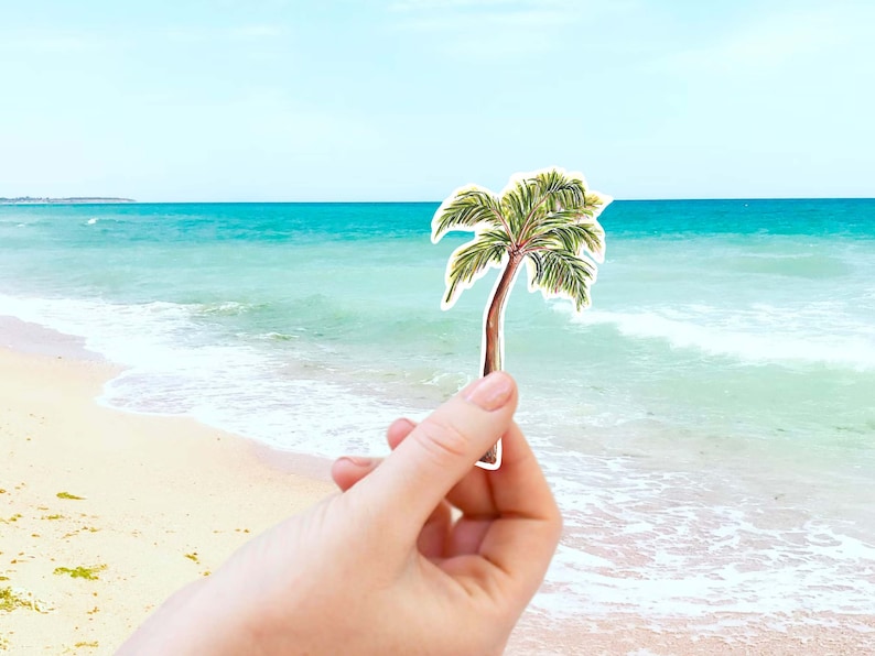 Puede incluir: Una mano sostiene un recorte de papel de una palmera verde sobre un fondo de oc&eacute;ano azul y playa de arena.