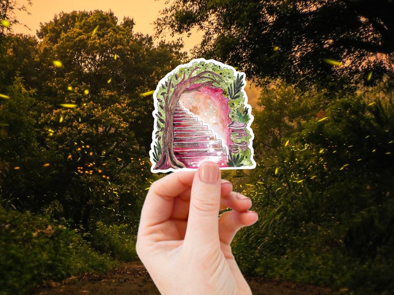 May include: A sticker featuring a watercolor illustration of a whimsical archway with stairs leading to a glowing light. The arch is framed by a tree and foliage, with a pink and red color scheme. The sticker is held in a hand against a blurred forest background.
