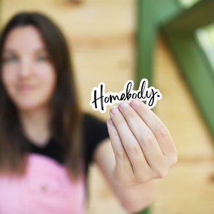 Pu&ograve; includere: Una persona tiene in mano un adesivo bianco con la parola "Homebody" scritta in corsivo nero.