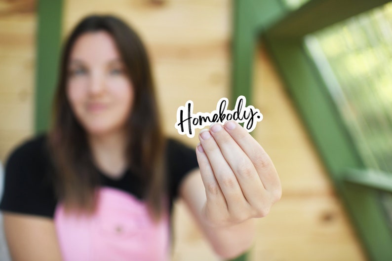Pu&ograve; includere: Una persona tiene in mano un adesivo bianco con la parola "Homebody" scritta in corsivo nero.