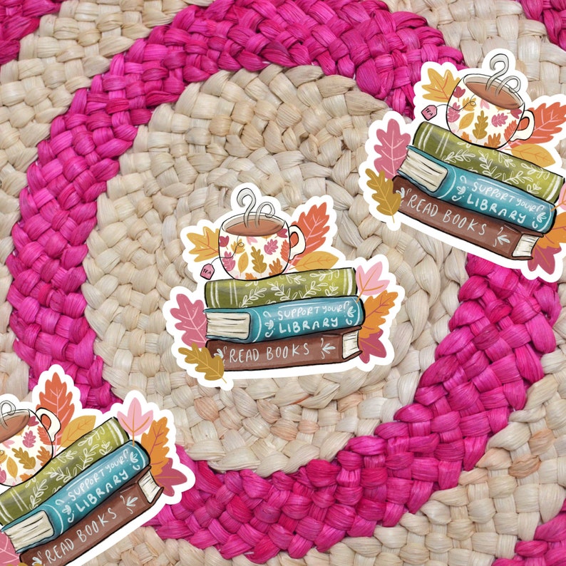 Op de afbeelding: Een sticker met een stapel boeken met de tekst "SUPPORT YOUR LIBRARY" en "READ BOOKS". Een kopje thee met een herfstontwerp staat bovenop de boeken. Herfstbladeren omringen de boeken en de kop, waardoor een gezellig, boekenachtig thema ontstaat.