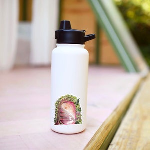 May include: A white insulated water bottle with a black cap and a whimsical sticker. The sticker features a watercolor illustration of a doorway framed by a tree with a staircase leading into a pinkish light.