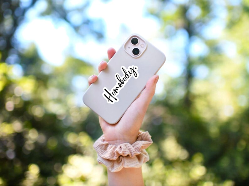 Pu&ograve; includere: Un iPhone bianco con un adesivo nero che dice "Homebody". Il telefono &egrave; tenuto in una mano con un elastico beige al polso.
