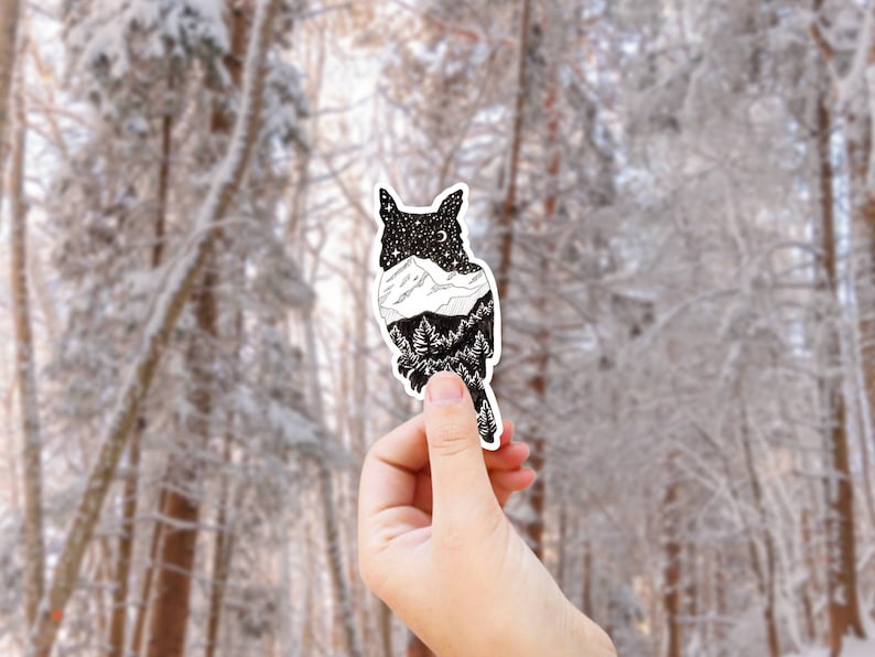 以下が含まれることがあります： 白黒のステッカー。体の内部に山脈と星が描かれたフクロウ。ステッカーは雪が積もった森のぼやけた背景を前に、手で持たれている。