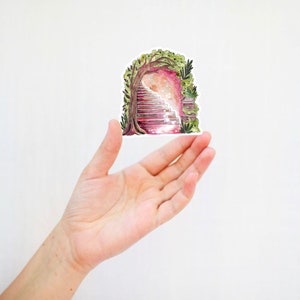 May include: A sticker featuring a watercolor illustration of a stone staircase leading into a glowing archway, framed by a tree and foliage. The sticker is held in a hand against a white background. The artwork has a fantasy theme.