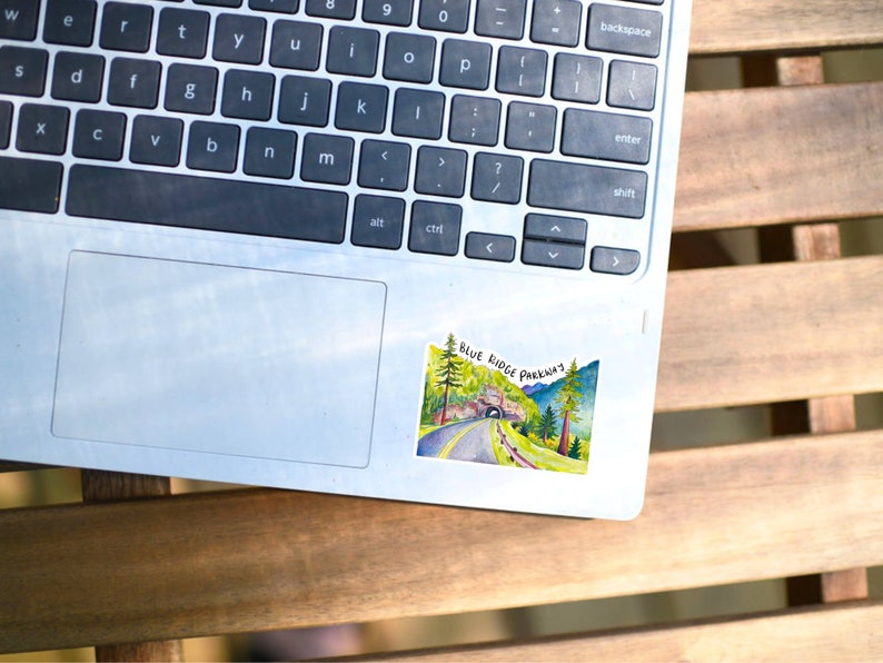 May include: A silver laptop computer with a sticker of a watercolor painting of a road through a tunnel with the text "BLUE RIDGE PARKWAY" on it.