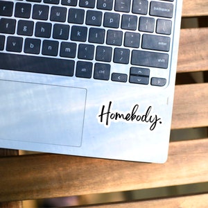 Pu&ograve; includere: Un computer portatile argentato con una tastiera nera e un trackpad. Il laptop ha un adesivo che dice "Homebody."