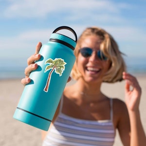 Puede incluir: Una botella de agua reutilizable azul con una pegatina de palmera en el lateral. La botella tiene una tapa negra y un asa de transporte negra.