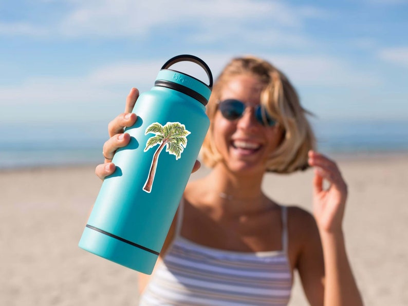 Puede incluir: Una botella de agua reutilizable azul con una pegatina de palmera en el lateral. La botella tiene una tapa negra y un asa de transporte negra.