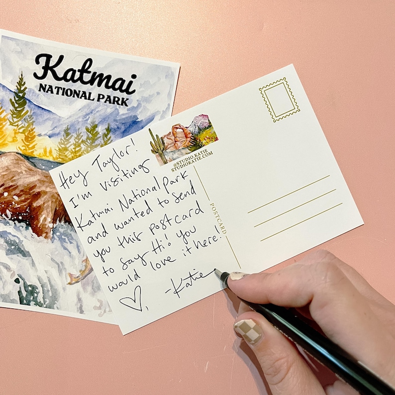 May include: A postcard with a watercolor illustration of a brown bear in a river. The postcard is addressed to Taylor and says "Hey Taylor! I'm visiting Katmai National Park and wanted to send you this postcard to say hi! You would love it here! -Katie"