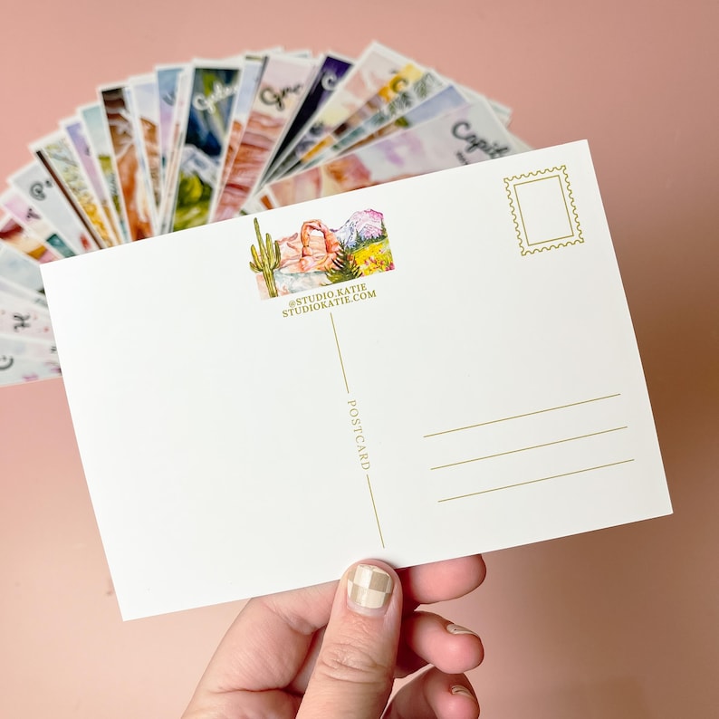 May include: A blank postcard with a watercolor illustration of a desert landscape with a cactus and mountains. The postcard has a gold line and the text "POSTCARD" printed on the back. The bottom of the postcard has the text "#STUDIO.KATIE STUDIOKATIE.COM".