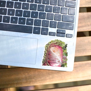 May include: A colorful sticker featuring a watercolor illustration of a staircase leading into a portal framed by a tree and foliage. The sticker is placed on a silver laptop, resting on a wooden table.