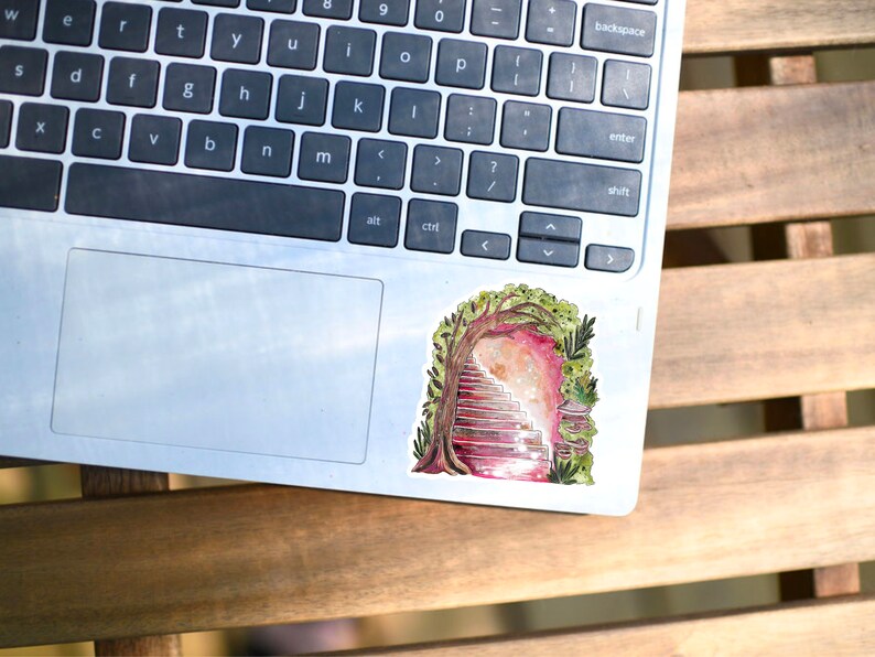 May include: A colorful sticker featuring a watercolor illustration of a staircase leading into a portal framed by a tree and foliage. The sticker is placed on a silver laptop, resting on a wooden table.
