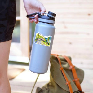 May include: A gray water bottle with a sticker on it. The sticker is a vintage postcard style image of a road with trees and the text "Nut Road Parkway".
