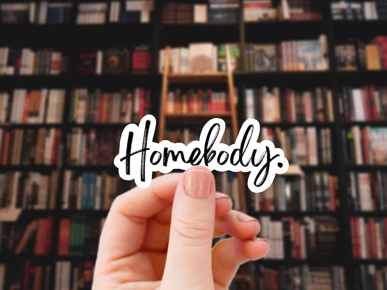 Pu&ograve; includere: Una mano tiene un adesivo bianco con la parola "Homebody" scritta in corsivo nero. L'adesivo &egrave; davanti a uno sfondo sfocato di librerie.
