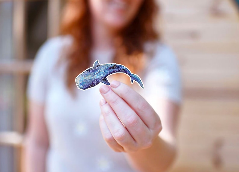 Op de afbeelding: Een blauwe en witte walvishaai-sticker met een gevlekt patroon. De sticker wordt in de hand van een persoon gehouden.