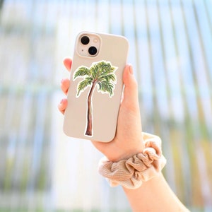 Puede incluir: Una funda de tel&eacute;fono blanca con una pegatina de palmera. La funda del tel&eacute;fono est&aacute; siendo sostenida por una mano que lleva una goma de pelo beige.