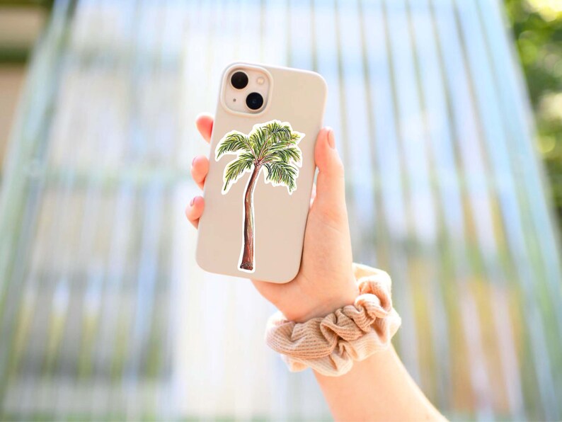 Puede incluir: Una funda de tel&eacute;fono blanca con una pegatina de palmera. La funda del tel&eacute;fono est&aacute; siendo sostenida por una mano que lleva una goma de pelo beige.