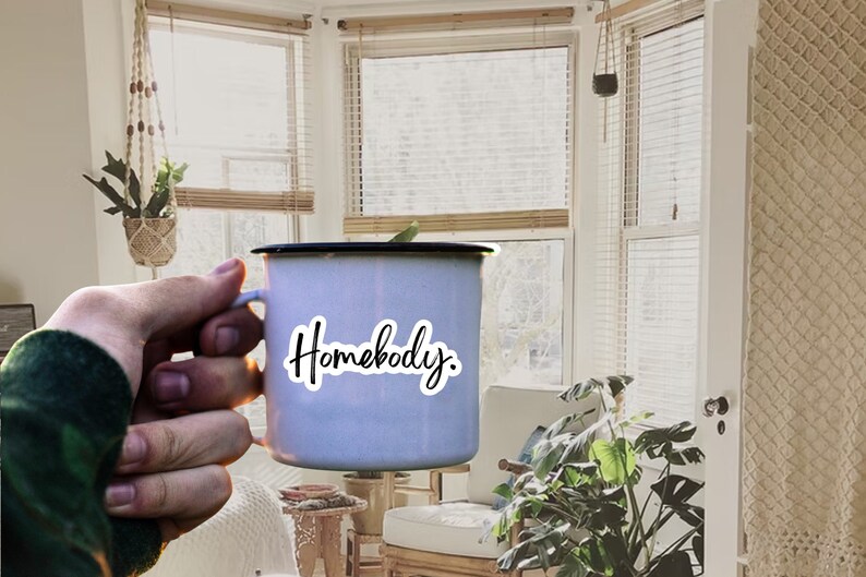 Pu&ograve; includere: Una tazza di smalto bianca con un bordo nero e un adesivo che dice "Homebody". La tazza &egrave; tenuta da una mano in un maglione verde.
