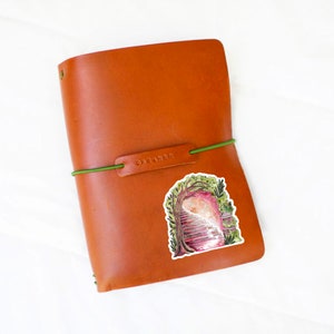 May include: A brown leather journal with a green elastic band and a sticker featuring a watercolor illustration of a staircase leading into a pink and white ethereal space. The word "DREAMER" is embossed on the leather.