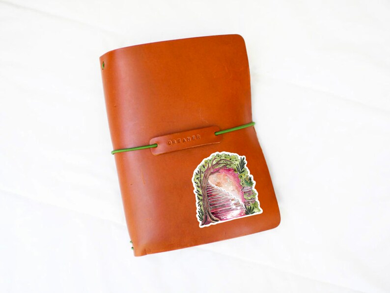 May include: A brown leather journal with a green elastic band and a sticker featuring a watercolor illustration of a staircase leading into a pink and white ethereal space. The word "DREAMER" is embossed on the leather.