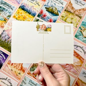 May include: A blank postcard with a watercolor illustration of Arches National Park in Utah. The postcard has a gold border and a stamp in the upper right corner. The text "#STUDIO KATIE STUDIOKATIE.COM" is printed in gold at the bottom of the illustration.
