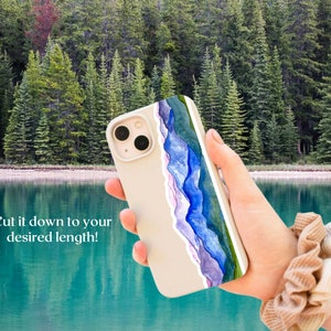 Pode incluir: Uma capa de telefone branca com um design de aquarela de montanhas azuis e roxas. A capa est&aacute; sendo segurada por uma m&atilde;o com um scrunchie marrom no pulso. O fundo &eacute; uma imagem desfocada de um lago e &aacute;rvores.