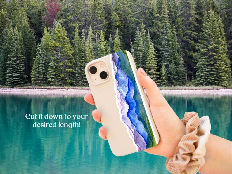 Pode incluir: Uma capa de telefone branca com um design de aquarela de montanhas azuis e roxas. A capa est&aacute; sendo segurada por uma m&atilde;o com um scrunchie marrom no pulso. O fundo &eacute; uma imagem desfocada de um lago e &aacute;rvores.