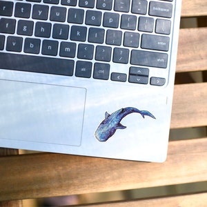Op de afbeelding: Een zilveren laptop met een blauwe en witte walvis sticker op het touchpad. De laptop staat op een houten oppervlak.