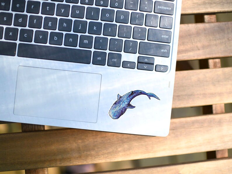 Op de afbeelding: Een zilveren laptop met een blauwe en witte walvis sticker op het touchpad. De laptop staat op een houten oppervlak.
