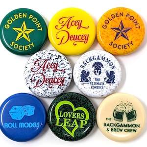 May include: A collection of colorful round buttons with various designs and text. The buttons feature phrases like "Golden Point Society," "Acey Deucey," "Roll Models," and "Lovers Leap." The buttons are on a white background.