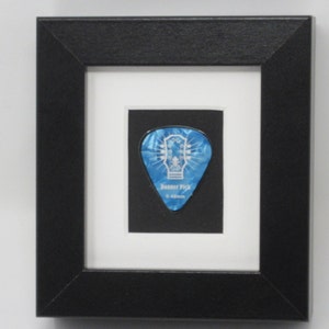 May include: A black picture frame with a white mat and a blue guitar pick inside. The pick has a white design and the text "Boomer Pick 0.48mm".