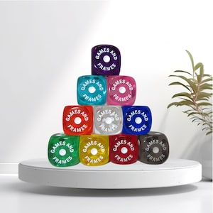 May include: A pyramid of ten colorful dice, each with the words "GAMES AND FRAMES" in white. The dice are various colors, including purple, teal, pink, red, clear, blue, green, yellow, maroon, and black. They are arranged on a white platform.