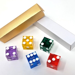 May include: Five translucent six-sided dice in purple, yellow, green, blue, and red, each featuring white pips. The blue die displays the number 444. Included are two rectangular storage boxes, one with a textured gold finish and one plain white.
