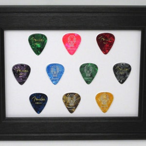 May include: A black framed display case with ten guitar picks inside. The picks are various colors and have different designs, including the Fender logo and the words "Heavy Pick".