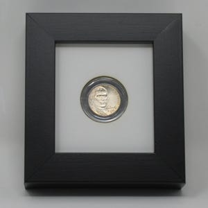 May include: A silver-colored United States five-cent coin featuring the profile of Thomas Jefferson, the text "Liberty" and "2022" encased in a circular protective capsule, mounted centrally within a square black frame with a white mat.