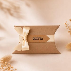 Op de afbeelding: Een bruine kartonnen kussendoosje met een crèmekleurig satijnen lint gestrikt tot een strik. Een rechthoekig bruin papieren label is bevestigd aan het midden van het lint, met de naam OLIVIA in zwarte hoofdletters op het label.
