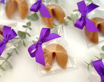 Puede incluir: Cinta morada atada alrededor de bolsas de celofán transparentes llenas de galletas de la fortuna. Las galletas de la fortuna son de color marrón dorado y tienen forma de media luna.