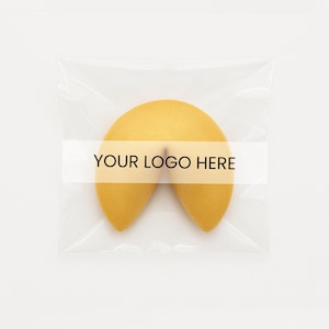 May include: A golden-yellow fortune cookie in a clear plastic wrapper. The cookie has a curved shape, with the text "YOUR LOGO HERE" printed on a white band across the center. The background is plain white.