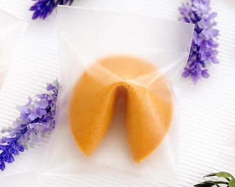 Puede incluir: Vista aérea de varias galletas de la fortuna en bolsas de plástico transparente, dispuestas sobre una superficie blanca. Ramas de lavanda púrpura y ramitas verdes se intercalan entre las galletas. Las galletas son de color marrón dorado.