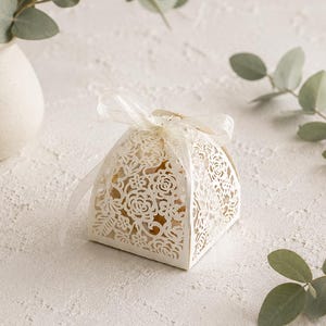 May include: A small, square-shaped favor box made of cream-colored cardstock with intricate laser-cut floral patterns on all sides. The box is tied at the top with a delicate, translucent white ribbon bow, creating a soft and elegant appearance.