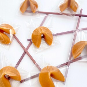 May include: An overhead shot shows several fortune cookies individually wrapped in clear plastic. The cookies are arranged on a white surface, with purple chopsticks forming a tic-tac-toe grid. The cookies are golden brown.