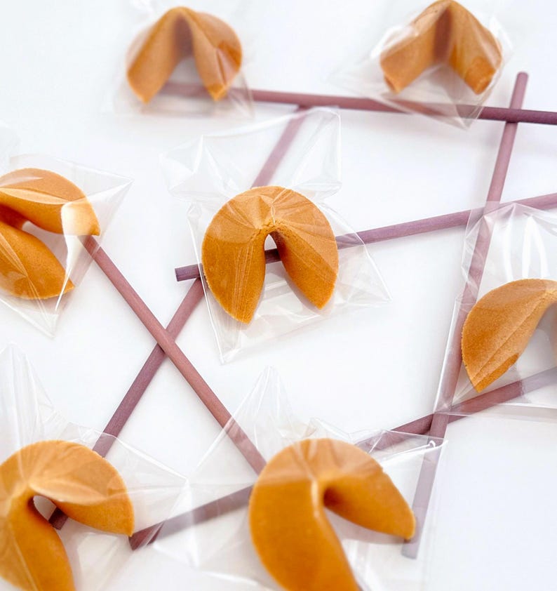 May include: An overhead shot shows several fortune cookies individually wrapped in clear plastic. The cookies are arranged on a white surface, with purple chopsticks forming a tic-tac-toe grid. The cookies are golden brown.