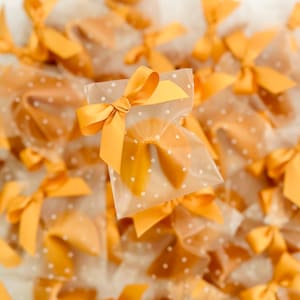 May include: A close-up of a pile of fortune cookies wrapped in clear cellophane bags with gold polka dots and tied with gold satin ribbons.