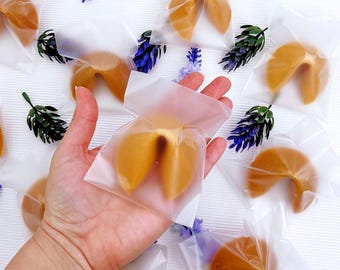 Puede incluir: Galletas de la fortuna doradas en bolsas de celofán transparente, dispuestas sobre una superficie blanca con ramitas de flores decorativas moradas y verdes. Una mano sostiene una galleta en una bolsa.