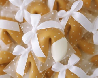 Puede incluir: Una colección de galletas de la fortuna de color marrón dorado, cada una envuelta individualmente en papel blanco translúcido con un patrón de pequeños puntos blancos. Cada galleta está atada con un lazo de satén blanco.