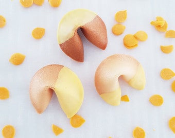 Puede incluir: Tres galletas de la fortuna con glaseado amarillo y marrón. Las galletas están sobre una superficie blanca con chispas amarillas.