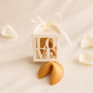 Op de afbeelding: Een klein wit kartonnen doosje met een uitgesneden venster waarin het woord LOVE staat, bovenaan gestrikt met een doorschijnend wit lint. Een goudbruin gelukskoekje ligt voor het doosje, dat ook een gelukskoekje bevat.
