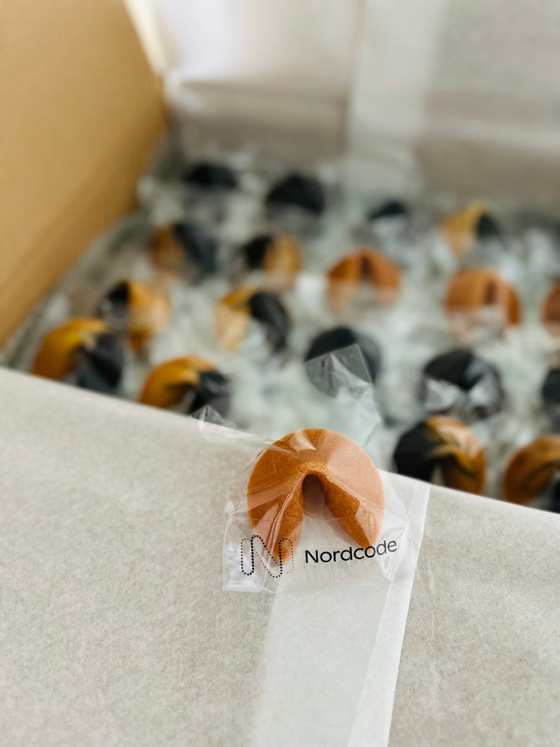 May include: A box of fortune cookies, some individually wrapped in clear plastic. One cookie is in focus, showing the "Nordcode" logo. The cookies are light brown and appear to be freshly baked, with a variety of colors.