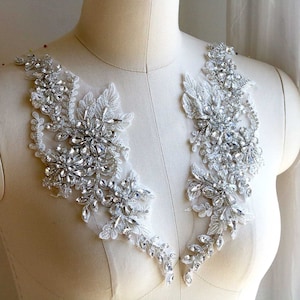 May include: Two white lace appliques with silver rhinestone embellishments. The appliques are shaped like a floral design and are perfect for adding a touch of sparkle to a wedding dress or other special occasion garment.